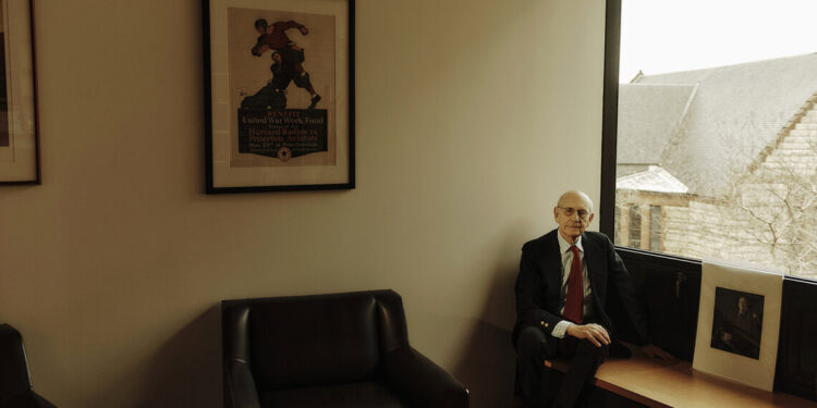 An older White man wearing glasses, a black suit, striped shirt and red tie, sits on a bench in the corner of his office.