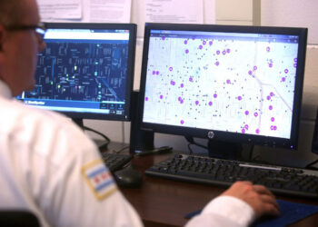 A person with light skin tone, wearing a white uniform with a Chicago flag on their shoulder, looks at gunshot detection programs on two computer screens.