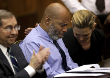 A Black man, wearing a blue striped button-down shirt and black tie, sits with an emotional look on his face and eyes closed. A White female lawyer, sitting on his right, is looking down and smiling while patting him on his back. A White male lawyer, sitting on his left, is smiling while clapping his hands.