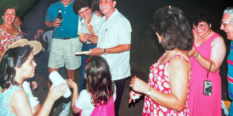 A middle-aged man wearing a white shirt and sunglasses laughs with family members during a party, in a photo from the 1980s.