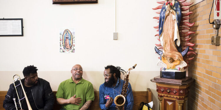 Three Black men sit in a chapel, next to a statue of Our Lady of Guadalupe. The man sitting in the middle wears glasses and a lime green polo over his dark green prison uniform. The two men sitting on either side of him hold a trombone and a saxophone, wearing jackets and slacks.