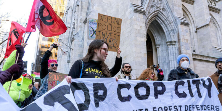 Several protesters hold a banner that reads "Stop Cop City," as they stand in front of a church. One protester in the background holds a sign that reads, "Defund the police. Invest in Black communities."