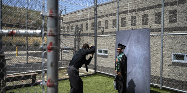 A photographer takes a picture of an incarcerated man, wearing a graduation cap, gown, a Mexican sarape print stole and a green stole that reads, "T.O.P.S.S." in front of a gray backdrop. The picture was taken outside on a lawn surrounded by barbed wire fences.