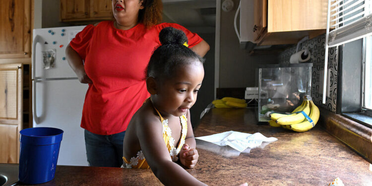 A young Black girl, wearing a yellow blouse, plays with seashells on a kitchen countertop. A Black woman, wearing a red blouse and red lipstick, stands with her hands on her hips in the background.