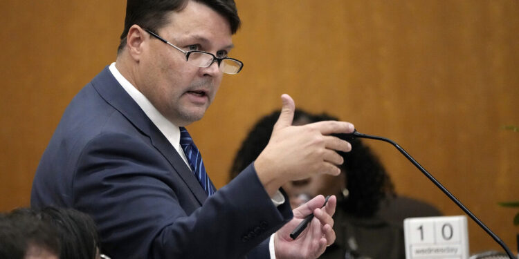 A White man, wearing glasses and a navy suit, speaks into a courtroom microphone.