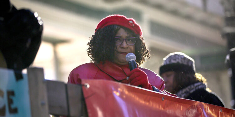 A Black female attorney wearing a red coat, hat and gloves, speaks into a mic during a protest.