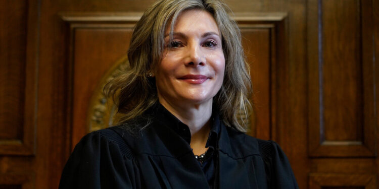 A White, blonde woman stands in a black robe behind a judge’s podium.