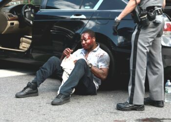 The badly beaten victim is seen sitting on the ground, leaning against a car after the attack.