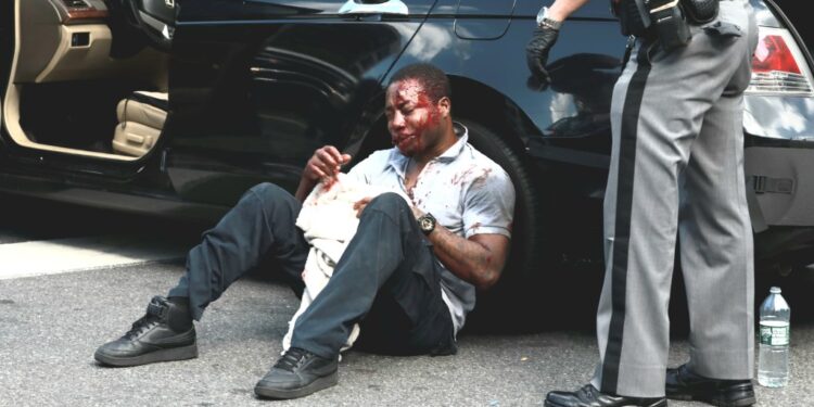 The badly beaten victim is seen sitting on the ground, leaning against a car after the attack.