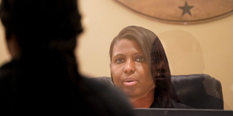 A Black woman with straight hair combed to the side sits at a judge’s bench behind a clear barrier. Another person’s reflection is visible on the barrier.