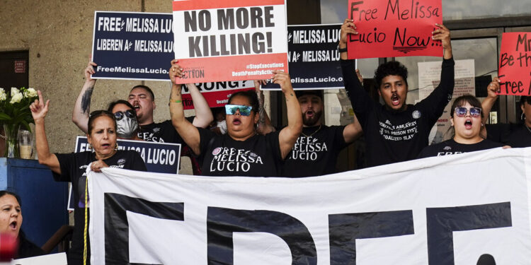 Outside of a building, a crowd representing mixed genders, ethnicities and ages stands holding signs above their heads with their mouths open. Some of the signs read “No more killing!” and “Free Melissa Lucio.”
