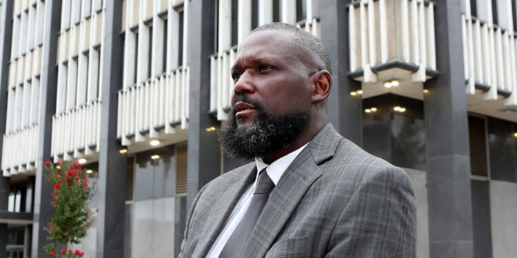 A Black male lawyer, wearing a gray suit, leans his arm on top of a wall for a portrait. A gray and white federal building is in the background.