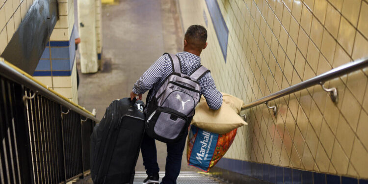 A man in a blue-checkered shirt descends a subway staircase lugging a large suitcase and a shopping bag.