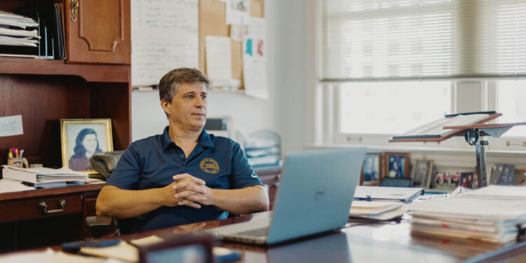 A White man, wearing a blue polo shirt, sitting in front of his office desk.