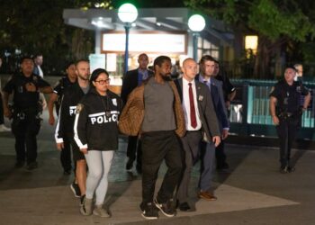 Derrick Mills in handcuffs is escorted by police officers near a subway station.