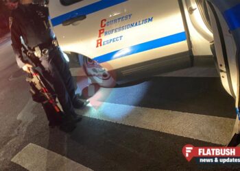 A cop holds the door open of a police cruiser while holding a black and red imitation assault rifle in the other hand.