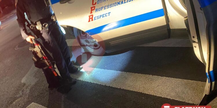 A cop holds the door open of a police cruiser while holding a black and red imitation assault rifle in the other hand.