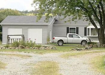 A suburban house with gray siding, black shutters, and two garage doors with a silver pickup truck parked out front and a large tree at the top of a gravel driveway.
