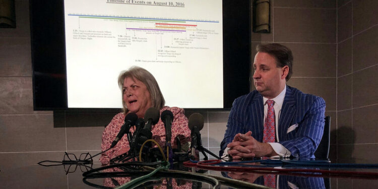 A White woman, wearing a red and pink patterned shirt, speaks into several news microphones inside a conference room. A male lawyer, in a red tie and blue pinstriped suit, sits next to her with his hands clasped on the table.