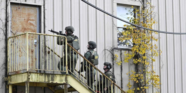 Three SWAT team members, wearing helmets, green uniforms and protective gear with the label "SHERIFF" on their backs, hold assault rifles while standing on a staircase.