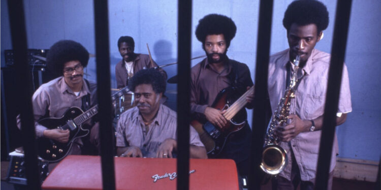 A musical band of Black men are playing instruments, such as a saxophone, guitar, piano, bass guitar and drums. They play behind bars in a room.