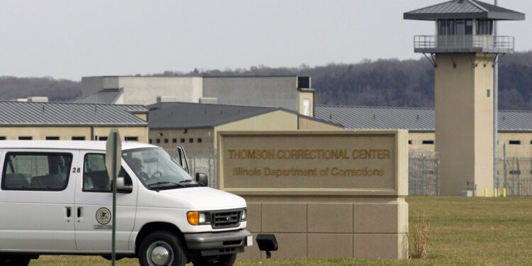A white van drives past a beige prison building with an observation tower and a sign out front that reads “Thomson Correctional Center, Illinois Department of Corrections.”
