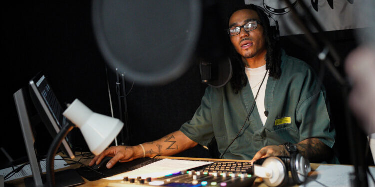 Darrius Turner, a Black man with glasses, works on radio equipment and wears headphones in a studio.