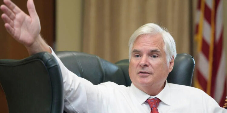 A White man with short white hair, wearing a white shirt and red tie, sits and holds his hand out towards the camera.