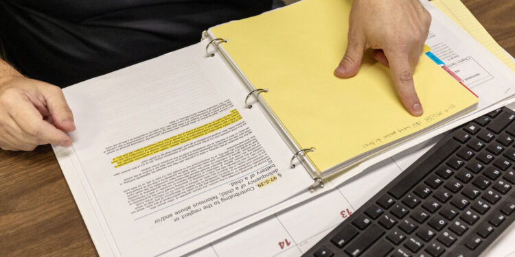 An image photographed from above of a man pointing to a yellow page in a binder, where he wrote down the criminal code containing the crime he believes women have committed when they use drugs while pregnant.