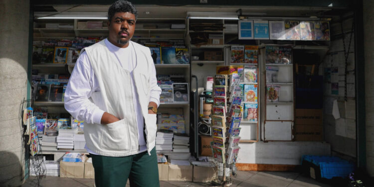 Donovan X. Ramsey, a Black man with short hair, wears a white shirt, vest and green pants, and stands in front of a newsstand.