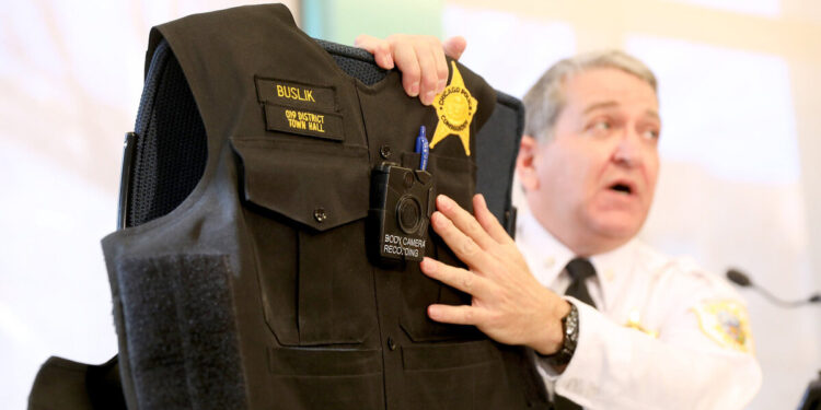 A White male police officer holds a police vest, pointing out its body camera.
