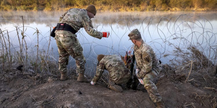 Three men in National Guard uniforms place razor wire alongside a river.