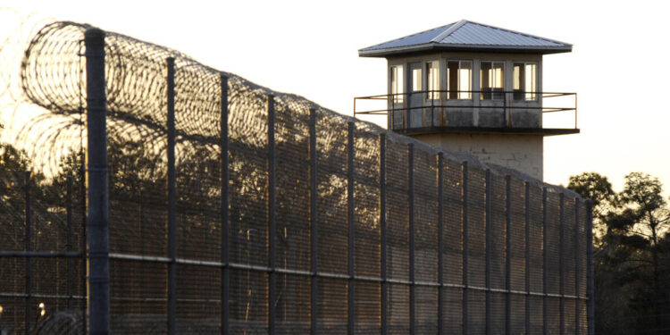 A photograph shows barbed wire and a prison tower at Holman Prison at sunset.