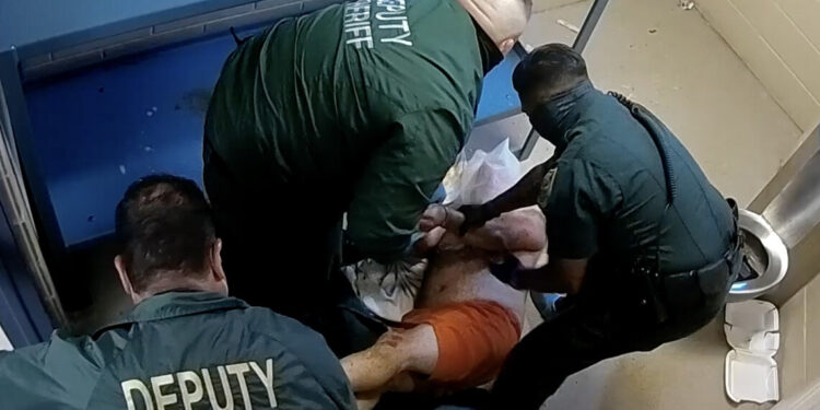 A man in orange prison shorts is being restrained by three officers in a prison cell.