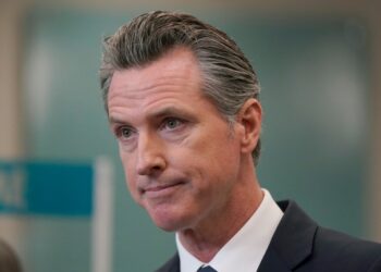 Governor Gavin Newsom speaking at a news conference, with a blurred background of an audience.