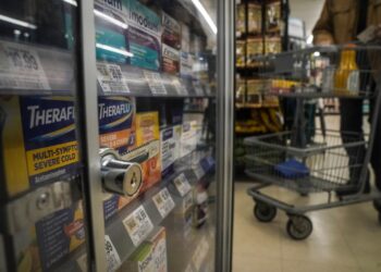 Locked pharmaceuticals in a glass cabinet at a store with a shopping cart visible in the background.