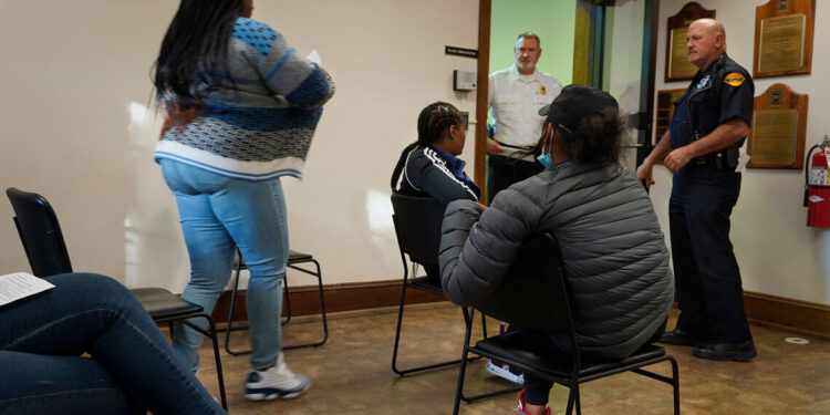 A Black woman walks out of a waiting room past three other Black women. Two White men in uniform stand at the front of the room by an open door.