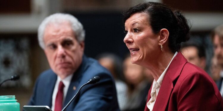 Colette Peters, a White woman in a red suit jacket and short brown hair, speaks, as Michael Horowitz, a White man in a navy suit jacket and white hair, looks on.