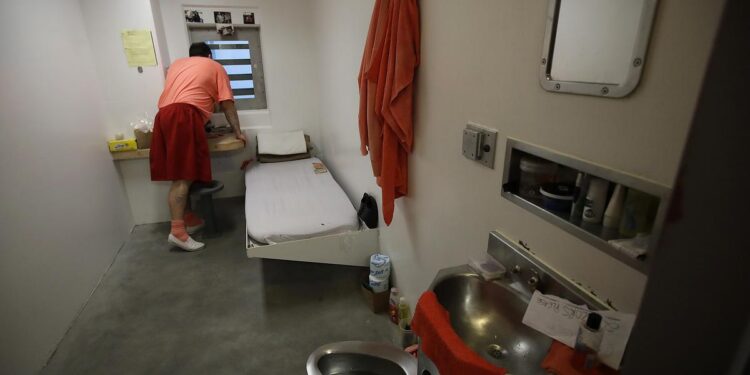 A man in an orange uniform looks out of a small window in his solitary confinement cell, which contains a narrow bed, a sink and attached toilet, and a small desk and chair.