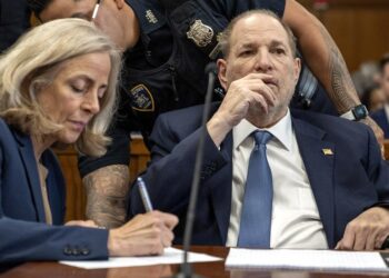 A White man, wearing a navy blue suit, is handcuffed to a chair as he sits next to his attorney in a courtroom. Two court officers stand behind them.