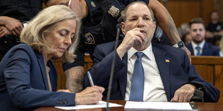 A White man, wearing a navy blue suit, is handcuffed to a chair as he sits next to his attorney in a courtroom. Two court officers stand behind them.