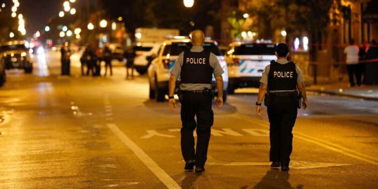 Two police officers walk in the middle of a street at night toward the scene of a shooting.