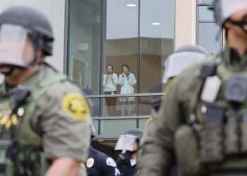 Two university members, wearing white tops, jeans and badges, look out a window and record videos from their phones. In the foreground, police wearing riot gear form a line in front of a building.