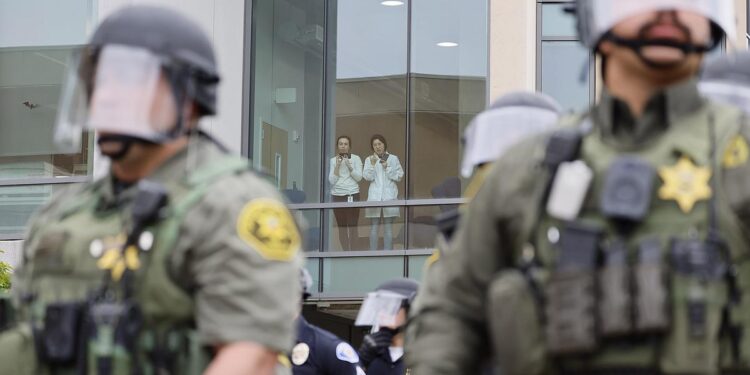 Two university members, wearing white tops, jeans and badges, look out a window and record videos from their phones. In the foreground, police wearing riot gear form a line in front of a building.