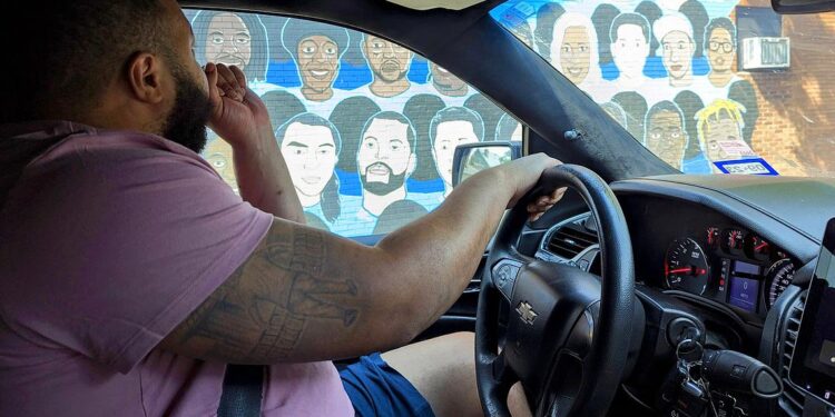 A Black man, wearing a light purple T-shirt and blue shorts, sits in the driver’s seat inside his car. He looks through his window at a mural of multiple portraits.