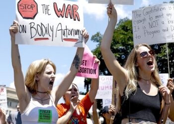 A White woman, wearing a white tank top and a label that reads "Forced motherhood is female enslavement," yells while holding a sign that reads, "Stop the war on bodily autonomy." People are protesting in the background.