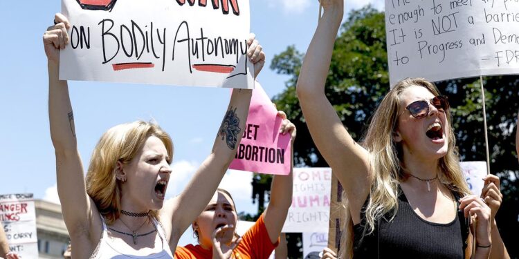 A White woman, wearing a white tank top and a label that reads "Forced motherhood is female enslavement," yells while holding a sign that reads, "Stop the war on bodily autonomy." People are protesting in the background.