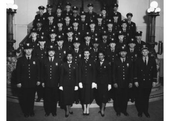 A black-and-white photo shows a group of uniformed police officers, including both men and women, standing in several rows on stairs. The officers are all wearing dark uniforms and caps, and they are posing for a formal group portrait inside a building.