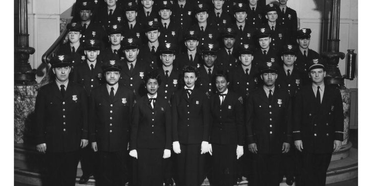 A black-and-white photo shows a group of uniformed police officers, including both men and women, standing in several rows on stairs. The officers are all wearing dark uniforms and caps, and they are posing for a formal group portrait inside a building.
