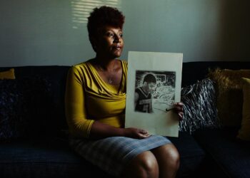 A woman wearing a yellow top and holding a drawing of her son playing basketball.
